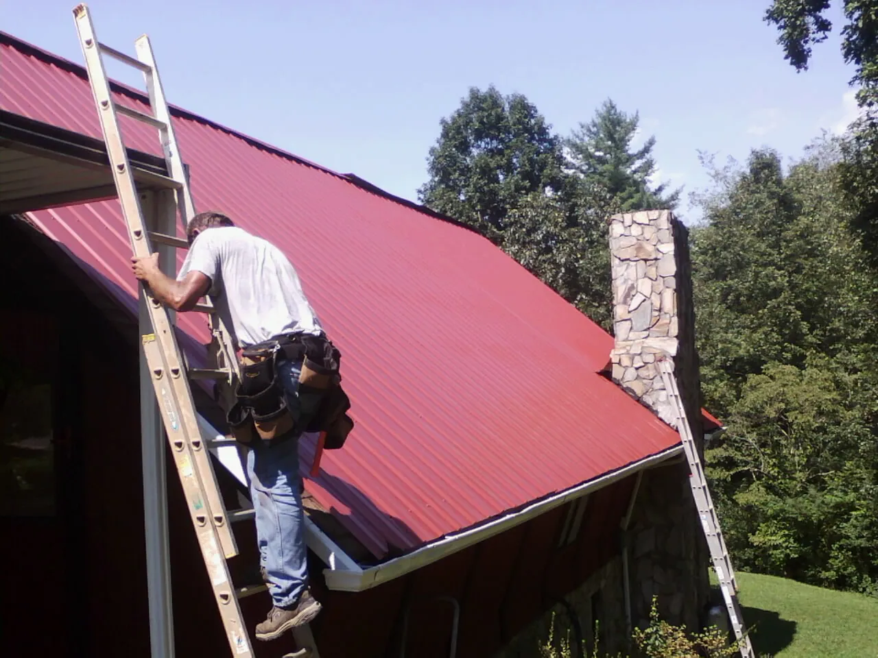 Roofer on a ladder beside a red metal roof and stone chimney — Soffit & Fascia Repair by Roofing Brothers in Clancy